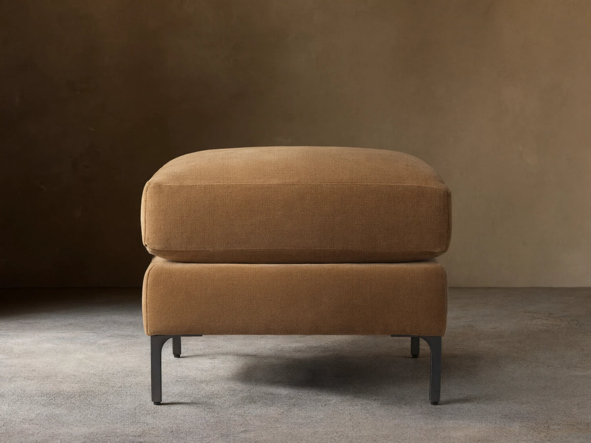 Sumner Ottoman in tan fabric on dark wooden floor, minimalist design, square shape, resting on slim black metal legs.