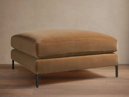 Sumner Ottoman with a plush, camel-colored fabric cushion and slender black metal legs, resting on a wooden floor with a neutral background.