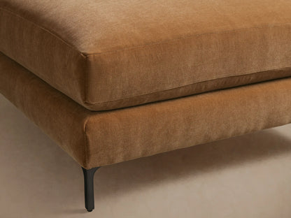 Sumner Ottoman in a warm brown tone, features a plush, square cushion and slim black legs. Placed on a wooden floor, against a neutral wall.