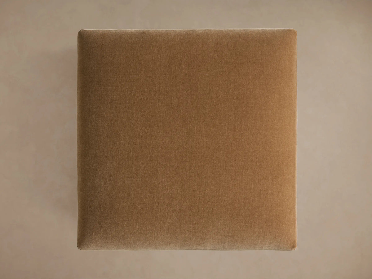 Sumner Ottoman in light brown fabric, close-up of plush cushion with square edges and metal legs, positioned against a neutral backdrop.