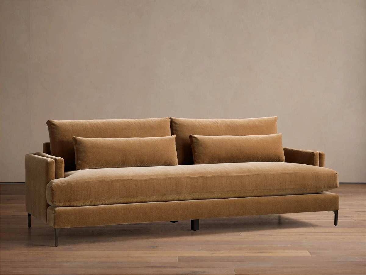 Front view of a caramel brown velvet sofa with two back cushions, two long lumbar pillows, and dark metal legs.