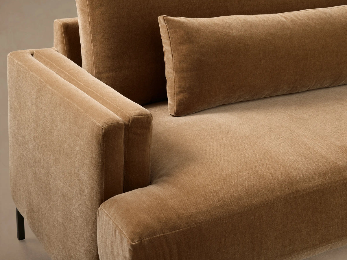 Front view of a warm brown velvet sofa with three back cushions, two lumbar pillows, and low square arms on dark legs.
