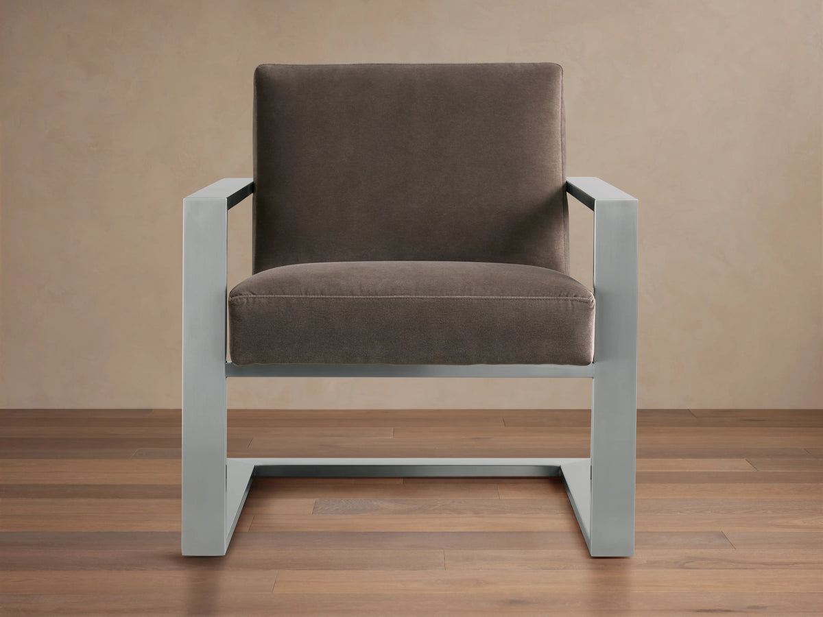 Sabina Chair with gray metal frame and brown cushioned seat and back, on wooden floor against a beige wall.