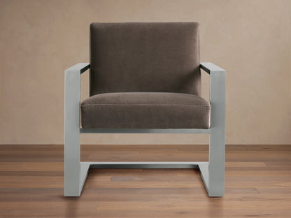 Sabina Chair with gray metal frame and brown cushioned seat and back, on wooden floor against a beige wall.