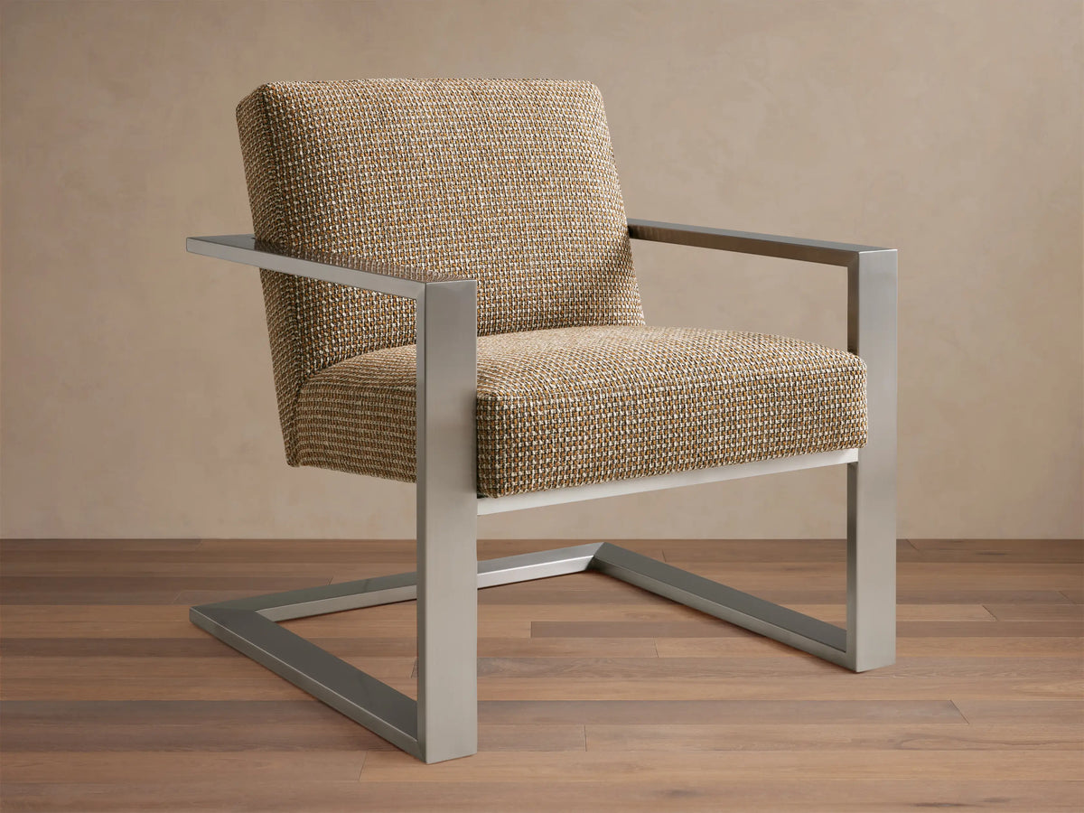 Sabina Chair with a textured beige upholstered seat and backrest, supported by sleek metallic angular legs on a wooden floor.