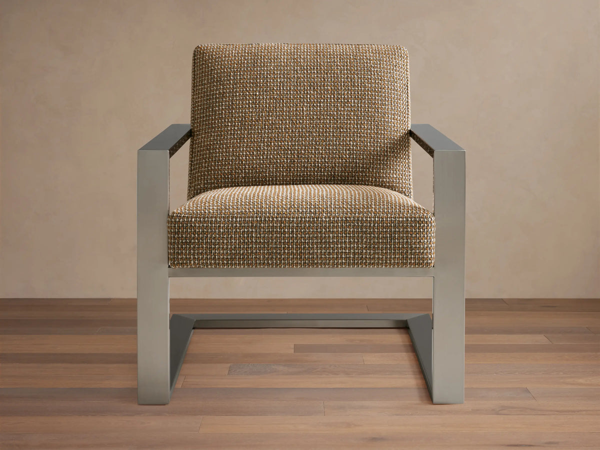 Sabina Chair with a textured beige upholstered seat and backrest, supported by sleek metallic angular legs on a wooden floor.