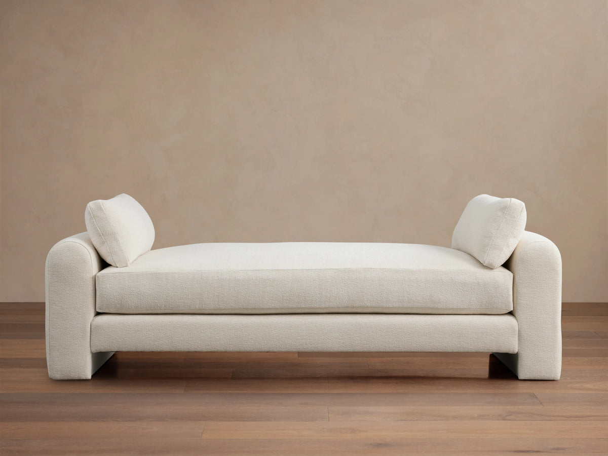 Beige Sloane Daybed on wooden floor, features plush cushions and soft fabric. Armrests and back cushions present.
