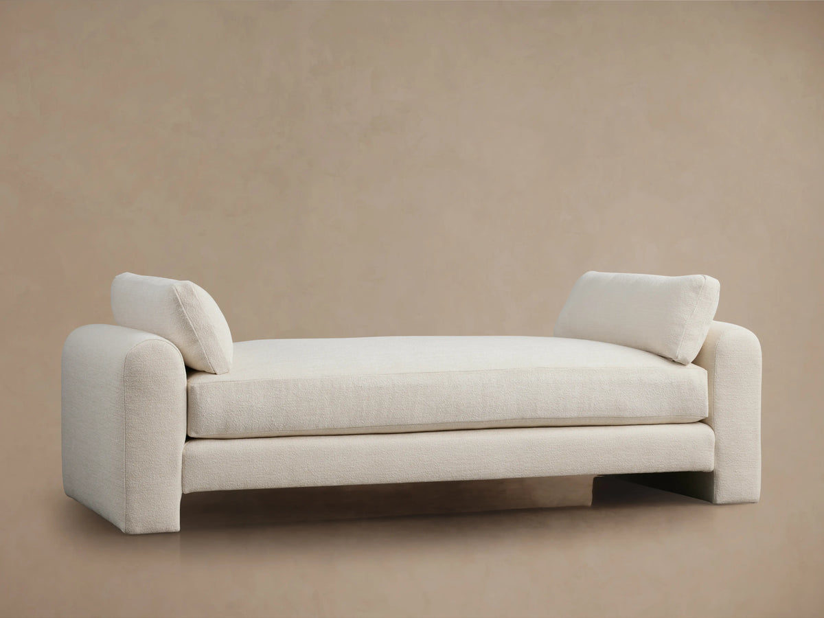 Beige Sloane Daybed on wooden floor, features plush cushions and soft fabric. Armrests and back cushions present.