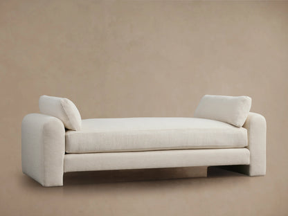 Beige Sloane Daybed on wooden floor, features plush cushions and soft fabric. Armrests and back cushions present.