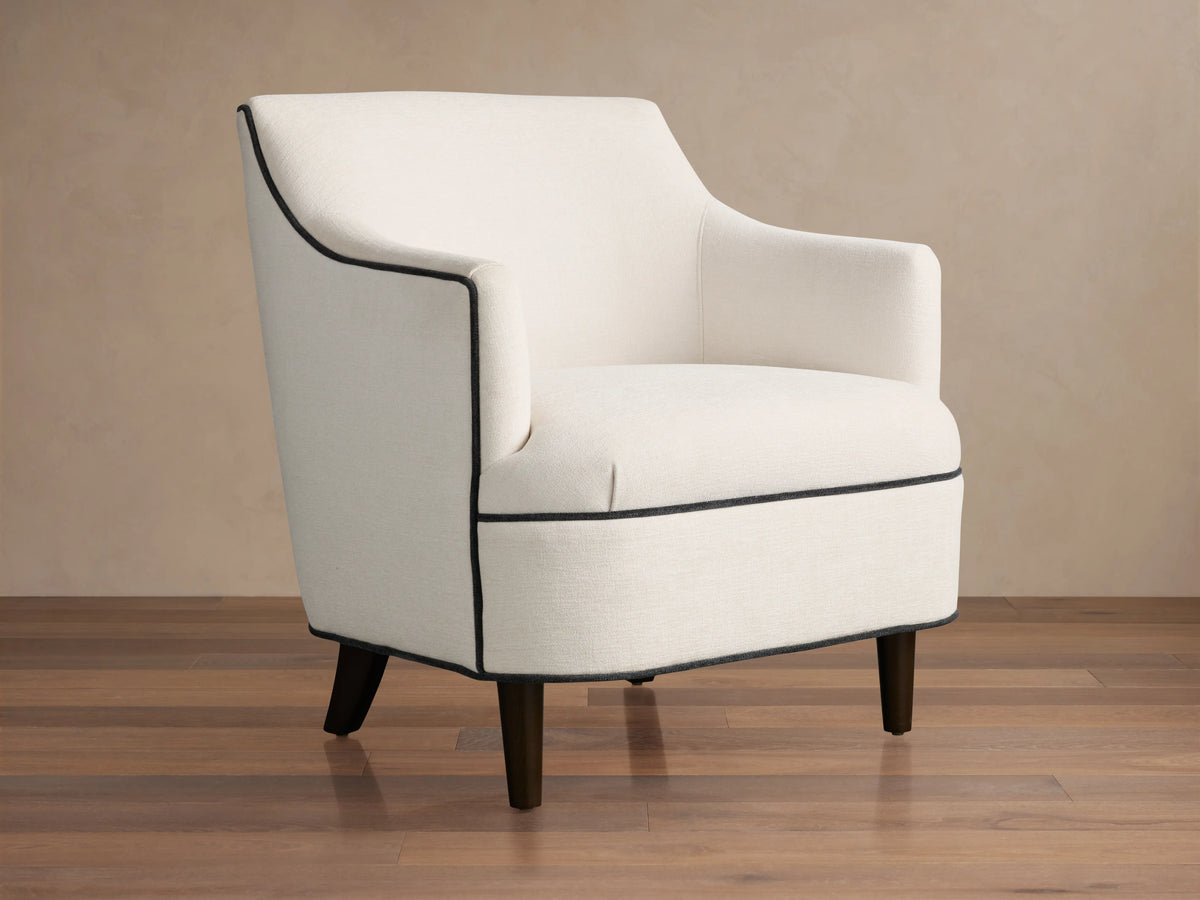 Savannah Chair: Cream upholstered armchair with black piping, dark tapered legs, angled on a wood floor.