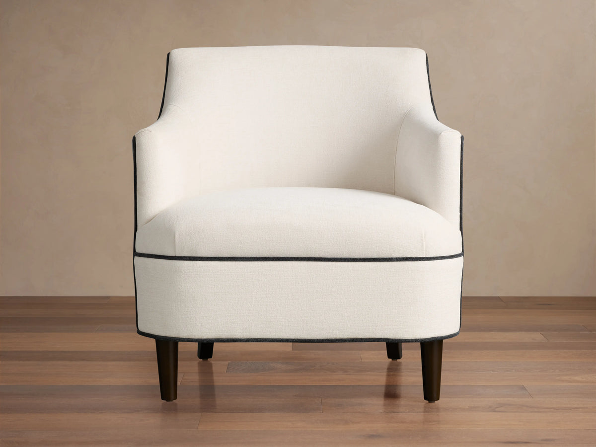 Savannah Chair: Cream upholstered armchair with black piping, dark tapered legs, angled on a wood floor.