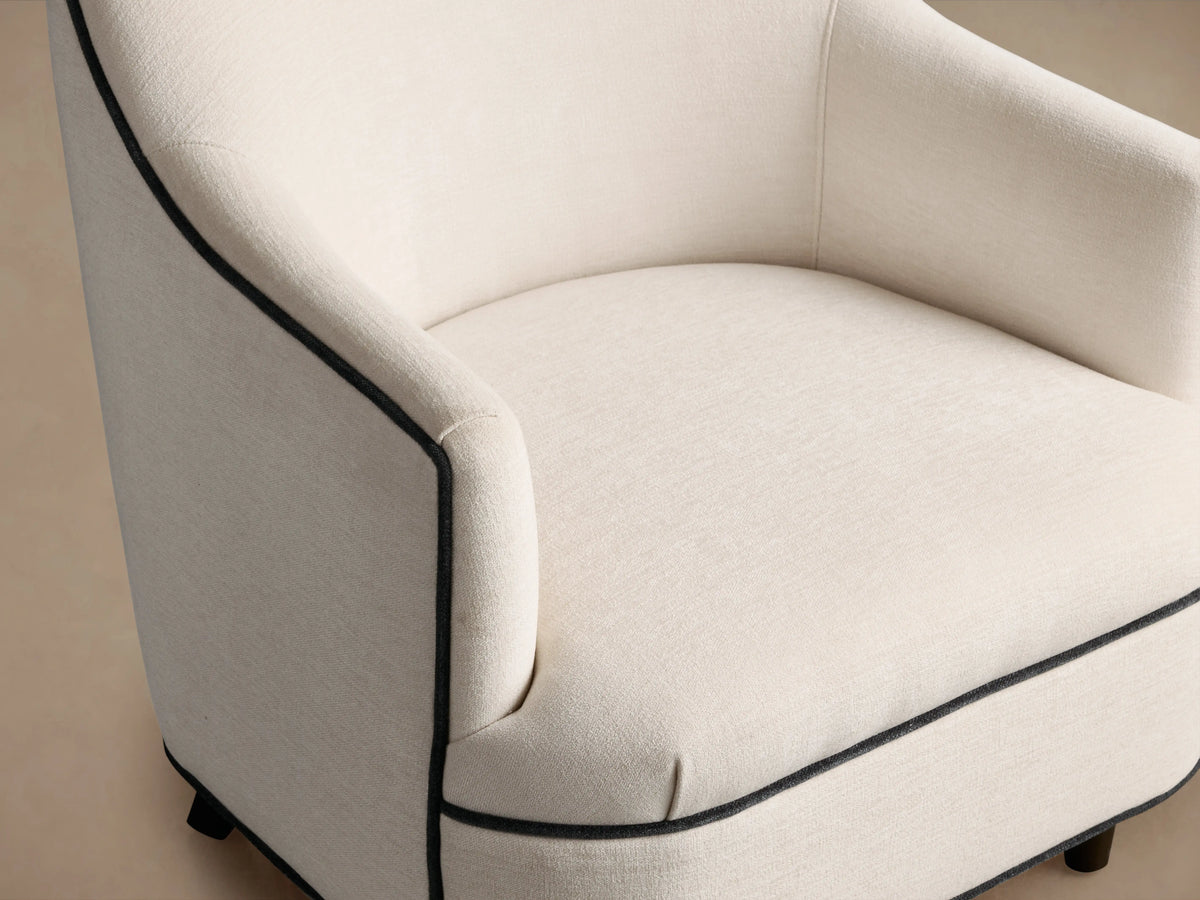 Cream Savannah Chair, upholstered with black piping along arms & base, dark wood tapered legs, on warm wood floor.