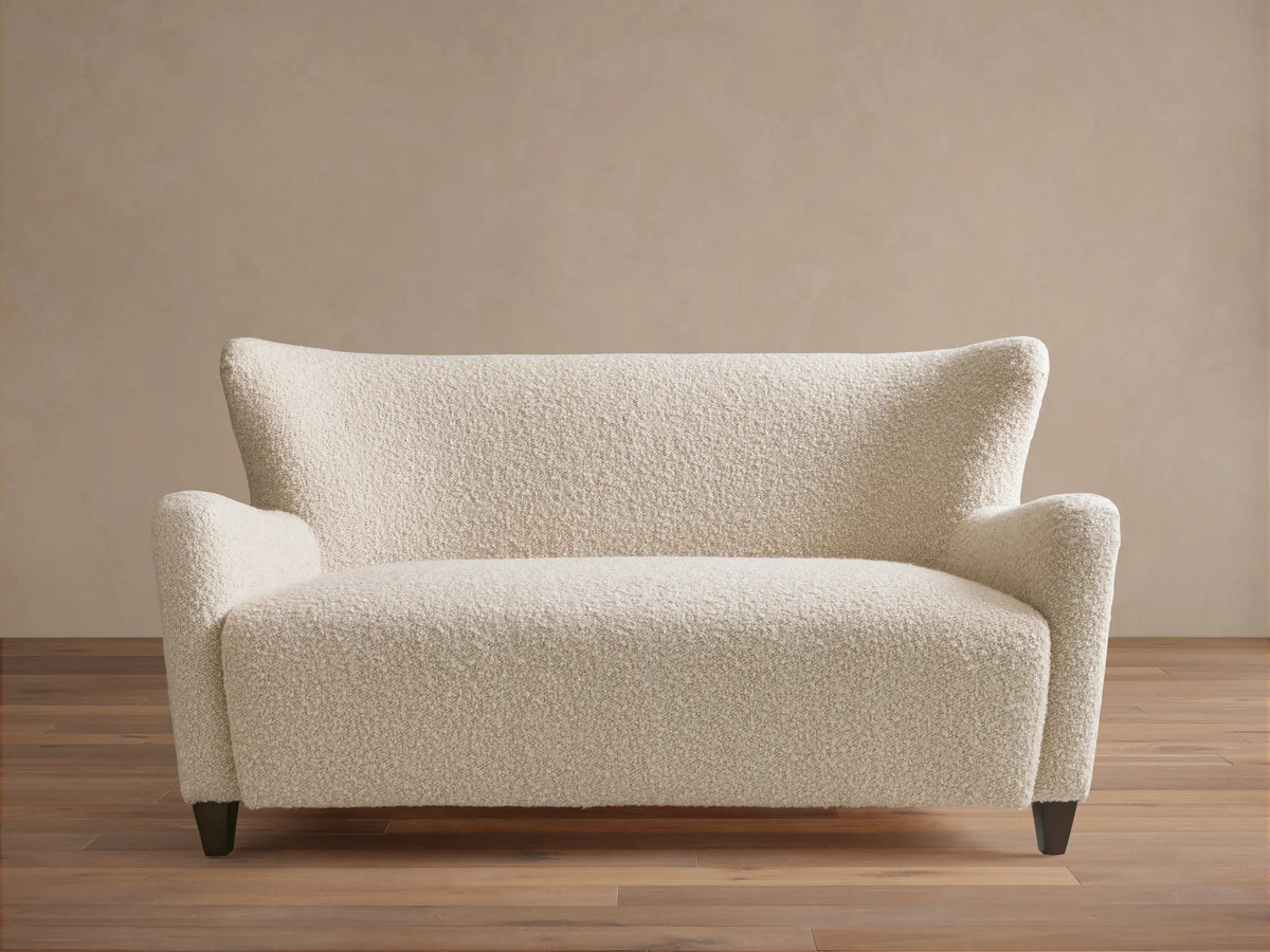 Sasha Settee in cream fabric featuring a textured, boucle finish, angled arms, and tapered dark wooden legs on a wooden floor.