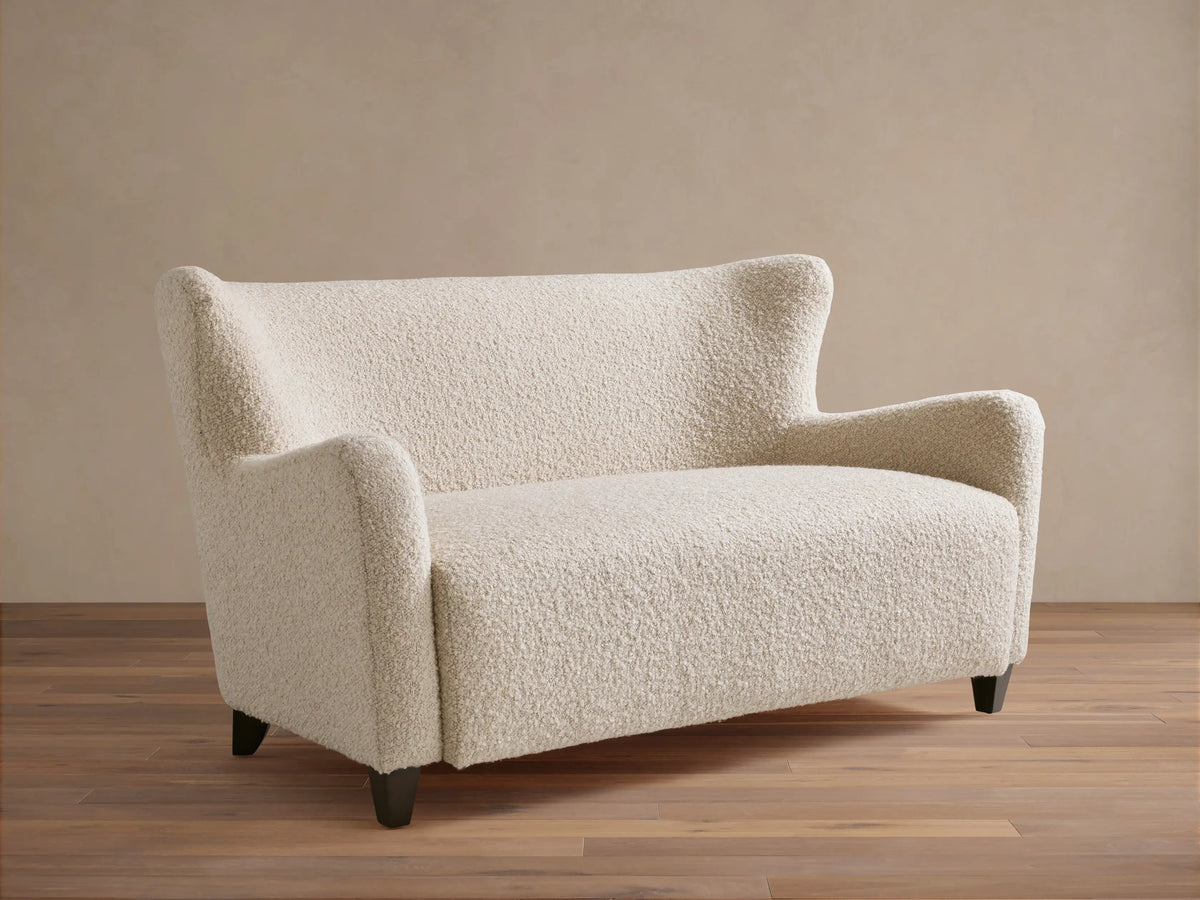 Sasha Settee in cream fabric featuring a textured, boucle finish, angled arms, and tapered dark wooden legs on a wooden floor.