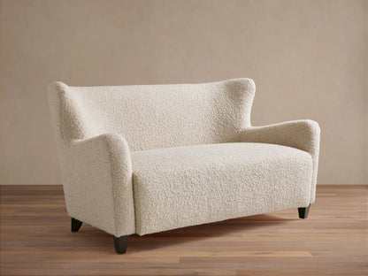Sasha Settee in cream fabric featuring a textured, boucle finish, angled arms, and tapered dark wooden legs on a wooden floor.