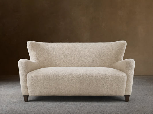 Sasha Settee in cream fabric featuring a textured, boucle finish, angled arms, and tapered dark wooden legs on a wooden floor.