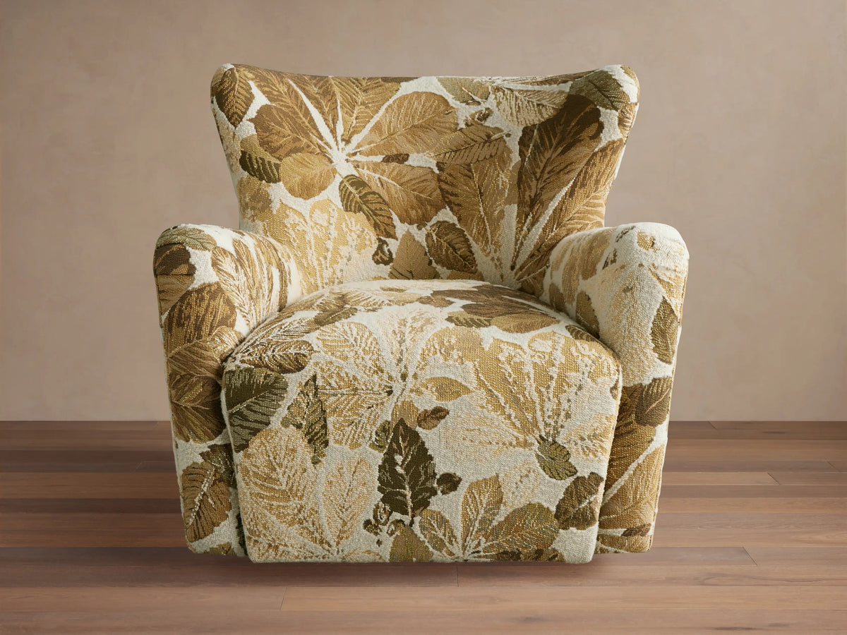 Swivel chair with high backrest and curved arms in a leaf-patterned fabric of green and brown tones on a wooden floor.