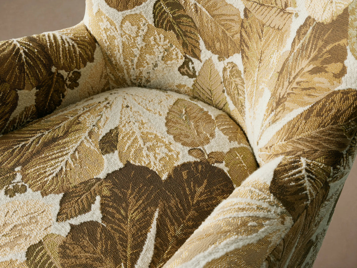Sasha Swivel Chair with a high backrest and wide armrests, upholstered in a leaf-patterned fabric with gold and olive tones, on wooden floor.