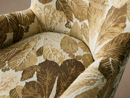 Sasha Swivel Chair with a high backrest and wide armrests, upholstered in a leaf-patterned fabric with gold and olive tones, on wooden floor.