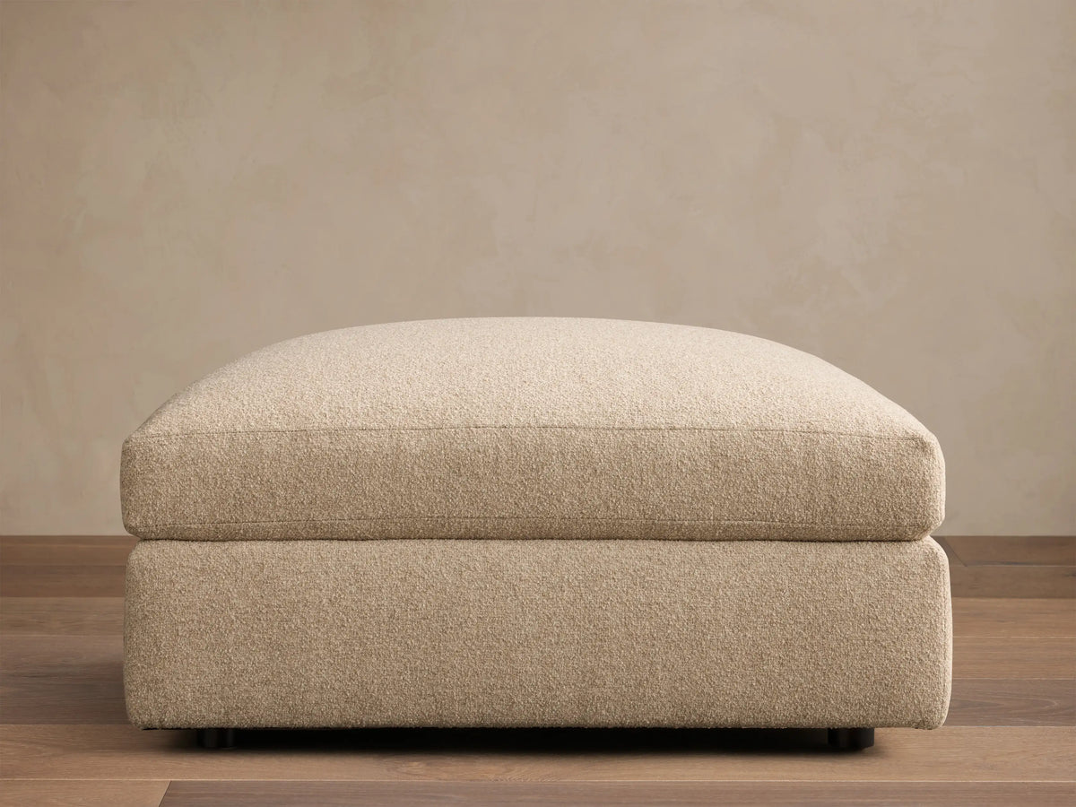 Front view of the light beige upholstered Stiles Ottoman, featuring a soft, rounded top cushion and blocky base.