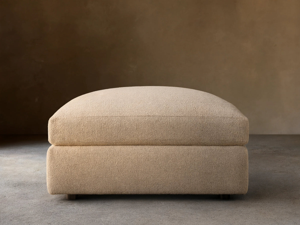 Front view of the light beige upholstered Stiles Ottoman, featuring a soft, rounded top cushion and blocky base.