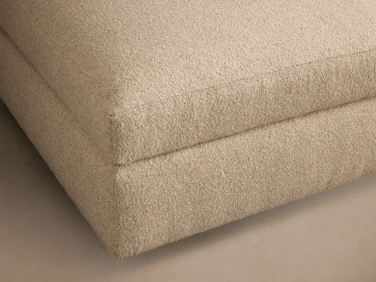 Front view of the light beige upholstered Stiles Ottoman, featuring a soft, rounded top cushion and blocky base.