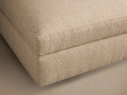 Front view of the light beige upholstered Stiles Ottoman, featuring a soft, rounded top cushion and blocky base.