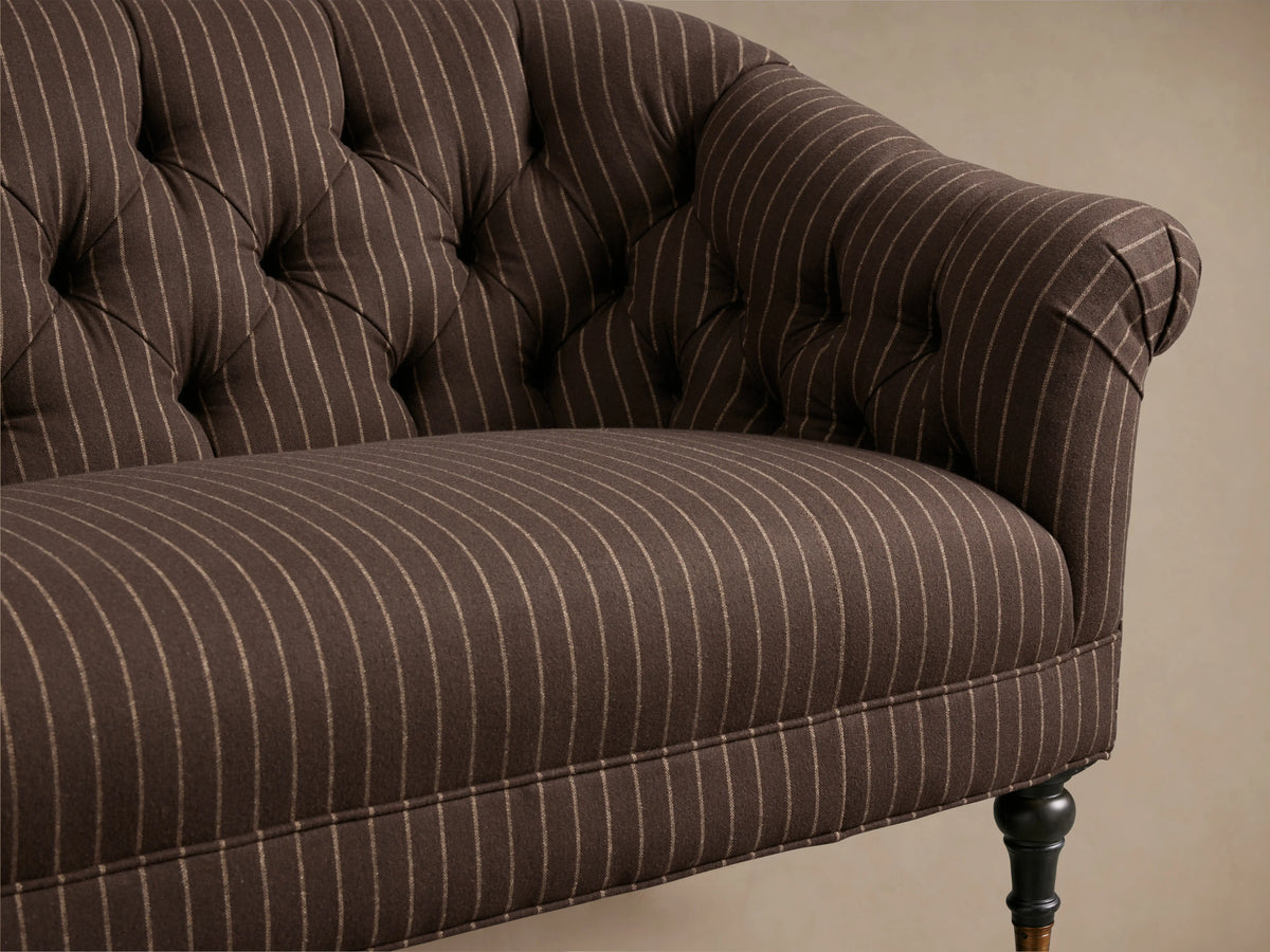 Tufted brown Tangier Settee with pinstripe pattern, curved arms, and dark turned wooden legs on casters, placed on wooden floor.