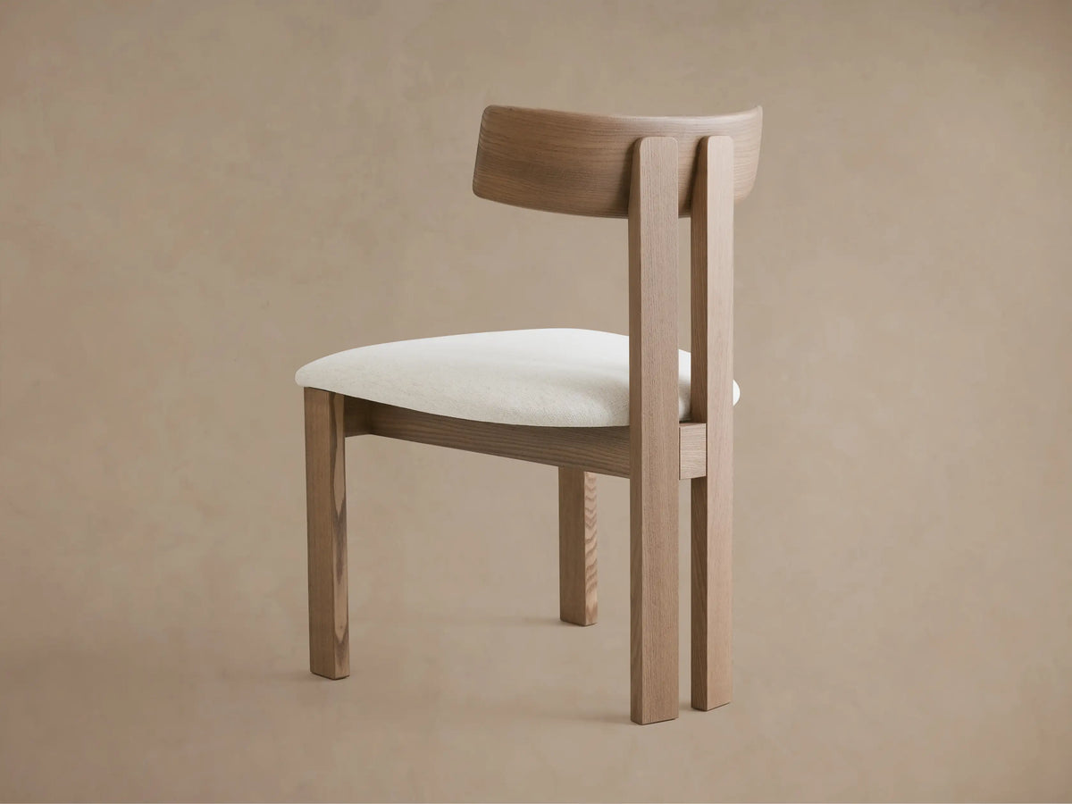 Rodin Wood Back Dining Chair. Light ash wood frame, curved backrest, and white textured fabric seat, angled view against beige wall.