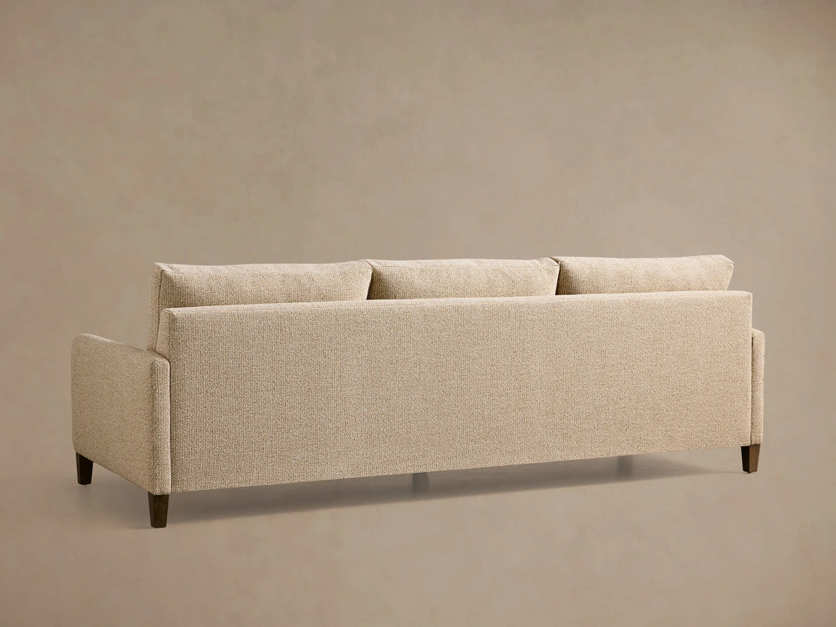 Rear view of a beige textured Kipton Slim Arm Elevated Three-Cushion Sofa with three back cushions and elevated mocha finish feet.