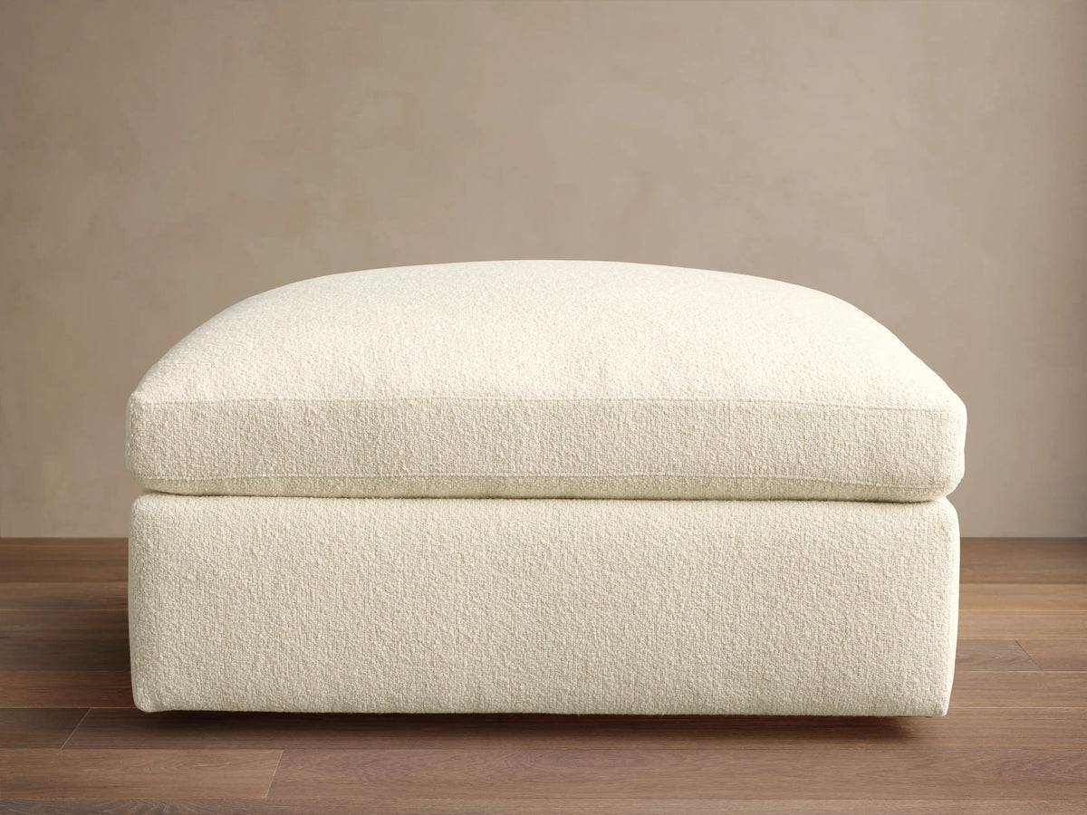 Textured cream Beale Ottoman with a plush, rounded top cushion, centered on a wooden floor against a plain beige wall.