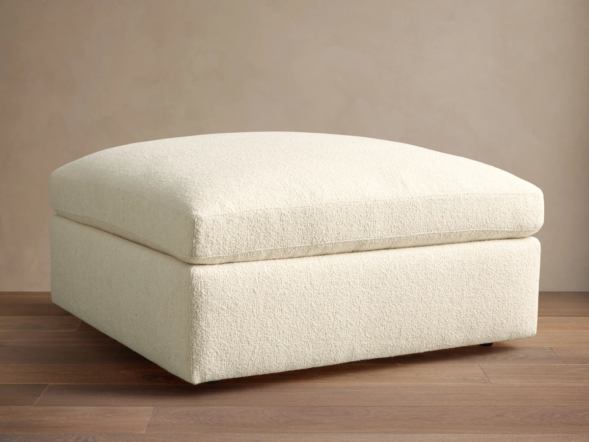 Textured cream Beale Ottoman with a plush, rounded top cushion, centered on a wooden floor against a plain beige wall.