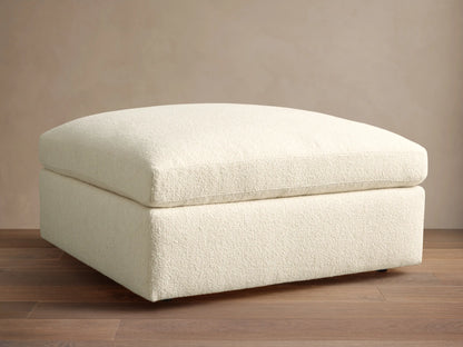 Textured cream Beale Ottoman with a plush, rounded top cushion, centered on a wooden floor against a plain beige wall.