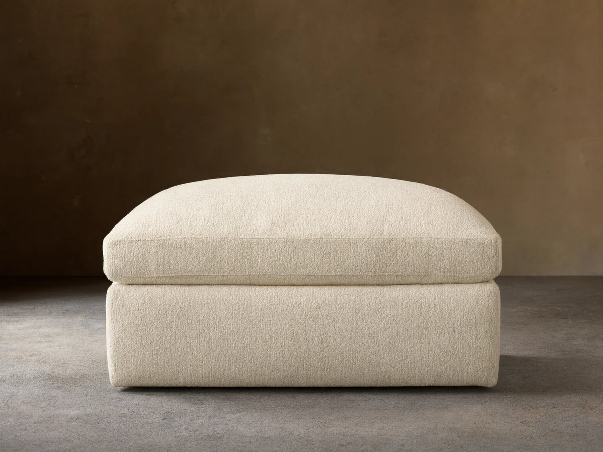 Textured cream Beale Ottoman with a plush, rounded top cushion, centered on a wooden floor against a plain beige wall.