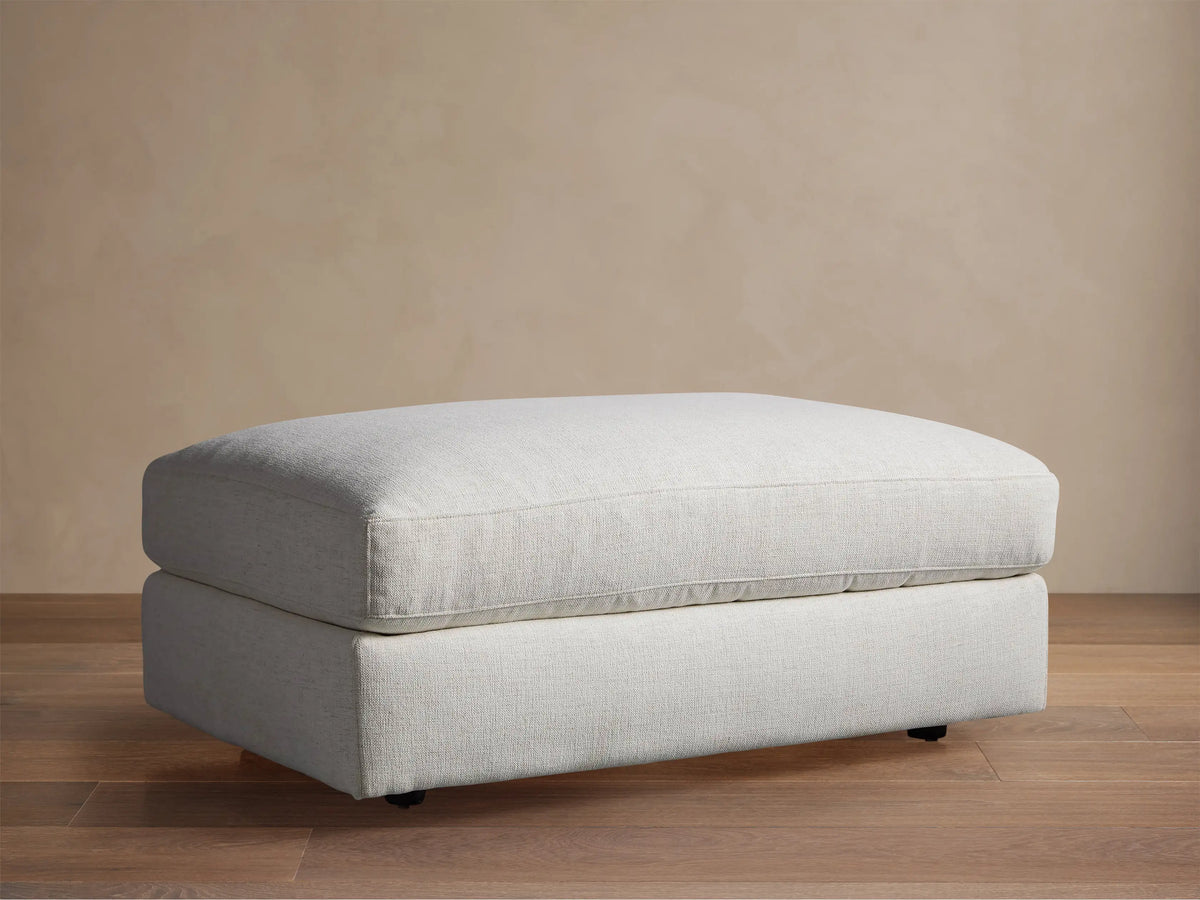 Light gray textured Beale Ottoman, rectangular with a plush top cushion, on wooden floor against a beige wall.