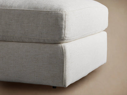 Beale Ottoman, a plush, light-toned upholstered ottoman with a soft, rounded cushion on a solid base, on a wood floor.