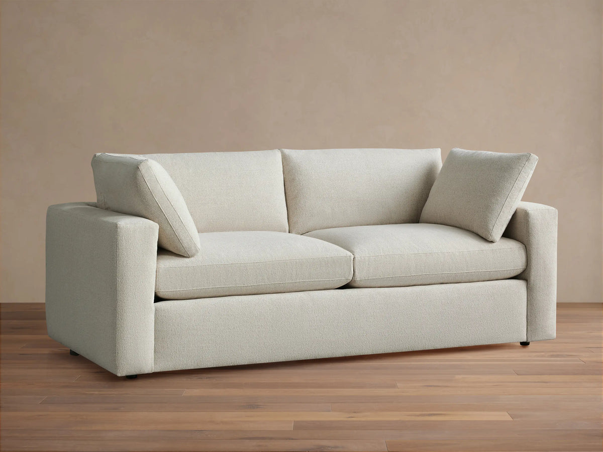 Front view of Beale Sofa in light cream textured fabric. Modern, casual design with wide arms, plush cushions, on wood floor.