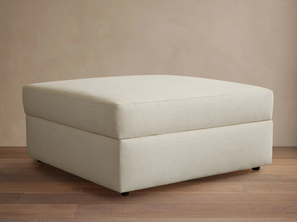 Front view of a rectangular cream ottoman with a visible textured fabric and dark small feet on a wooden floor.