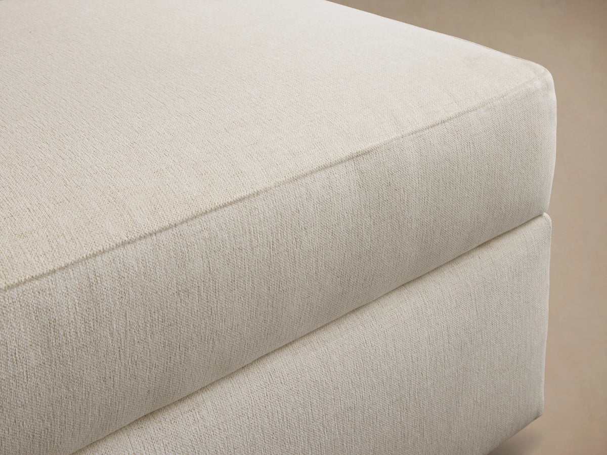 Front angled view of a cream upholstered square ottoman with a thick, plush top cushion and low-profile dark feet.