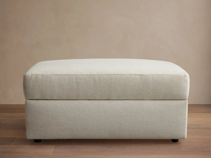 Front view of a light beige upholstered ottoman with a rectangular shape and a structured top cushion.