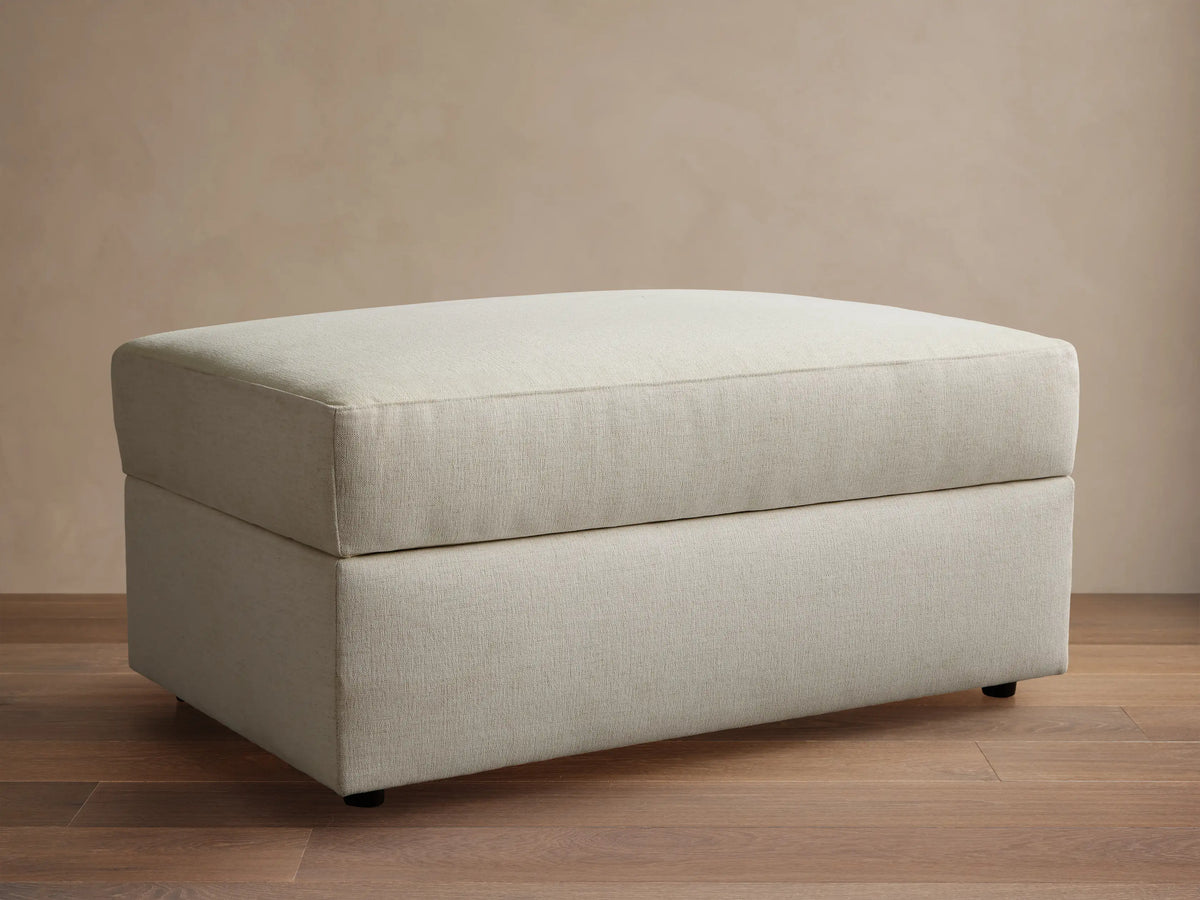 Front view of a light beige upholstered ottoman with a rectangular shape and a structured top cushion.