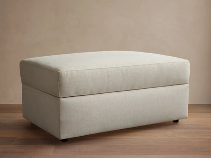 Front view of a light beige upholstered ottoman with a rectangular shape and a structured top cushion.