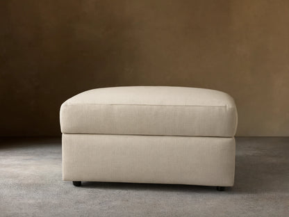 Front view of a light beige upholstered ottoman with a rectangular shape and a structured top cushion.